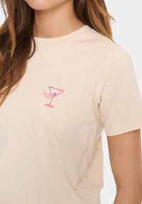 Béžové bavlněné tričko s růžovým potiskem martini sklenice a textem "chin chin" umístěným na levé části hrudi.