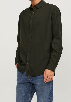 Hombre vestido con una camisa de manga larga verde oscuro abotonada y con cuello, y vaqueros azules, de pie con una mano en el bolsillo.
