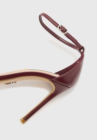 Sandale à talons hauts bordeaux avec une tige en cuir brillant, une sangle au niveau de la cheville avec une boucle métallique et une semelle intérieure beige.