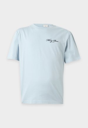 Jeune homme portant des lunettes de soleil foncées, un t-shirt bleu clair à manches courtes avec un texte, un jean bleu ample et des baskets noires, debout avec une main dans la poche.