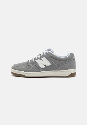 Zapatilla deportiva de ante gris con el logo "N" blanco, cordones blancos y suela marrón claro, mostrada sobre un fondo blanco, vista lateral.