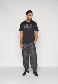 Svart Adidas t-skjorte med stor logo, kombinert med grå joggebukser med mønster og svarte treningssko. Enkel design og avslappet passform.