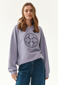 Felpa viola chiaro realizzata in tessuto morbido, con un grande logo circolare testurizzato sul davanti. Abbinata a jeans in denim blu.