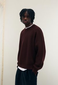 Bordeaux-röd stickad tröja med ribbat mönster, rundad halsringning och oversized passform, bärs över en vit skjorta och matchas med mörka manchesterbyxor.