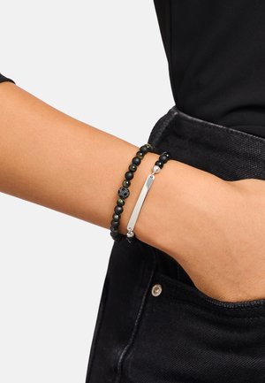 Polso con due braccialetti di perline, uno con perline nere e verdi e una perlina testurizzata, l'altro con una barretta d'argento, indossati sopra jeans neri.