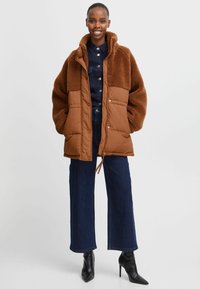 Brun polstret jakke med en fluffy sherpa-overdel, der har store frontlommer og en snor i kanten, parret med mørke denim og højhælede støvler.