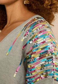 Szürke pulóver színes flitterekkel a vállán. A flitterek különböző árnyalatokban, csíkokban rendezve, textúrát és vizuális érdeklődést teremtenek.