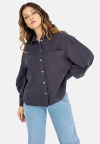 Ciemnoszara koszula z guzikami, posiadająca dwie przednie kieszenie, luźny krój, długie rękawy oraz teksturę materiału. W zestawie z jasnoniebieskimi dżinsami.