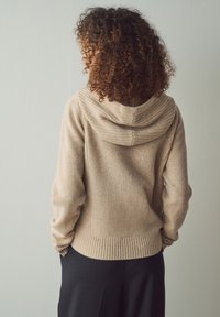 Maglione beige a maglia con cappuccio, dettagli a coste sul collo e sull'orlo, realizzato in materiale morbido, indossato sopra pantaloni scuri.