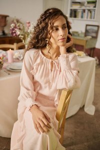 Žena s kovrčavom kosom u svijetloružičastoj haljini sjedi na drvenoj stolici za ukrašenim stolom za blagovanje s cvijećem i upaljenom svijećom.