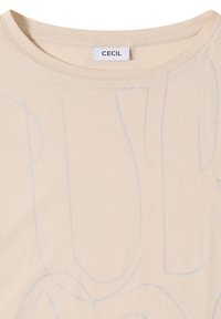 T-shirt beige à col rond avec de grandes lettres discrètes en gris clair contours, et une étiquette blanche portant la mention "CECIL" cousue à l'intérieur du col.