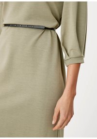Zielona oliwkowa sukienka z długimi rękawami, z zaakcentowaną talią podkreśloną cienkim czarnym paskiem z metalowymi ćwiekami. Gładki, fakturowany materiał.