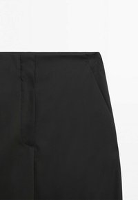 Gros plan sur la ceinture et la couture de la poche latérale d'un vêtement noir avec un tissu lisse sur un fond blanc uni.