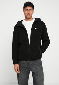 Mustavilla fleece-tyylillä varustettu vetoketjuhuppari, jossa on teksturoitu pinta, kontrastiväriset mustat reunat, etutaskut ja pieni logopatchi rinnassa.