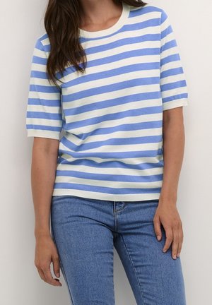 Femme portant un pull à manches courtes rayé horizontalement bleu et blanc et un jean en denim bleu, debout contre un mur clair uni.