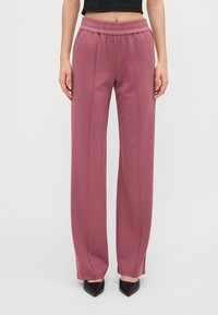 Roze, wijde broek met elastische tailleband. Gladde stof met twee verticale naden aan de voorkant, gecombineerd met zwarte puntige hakken.