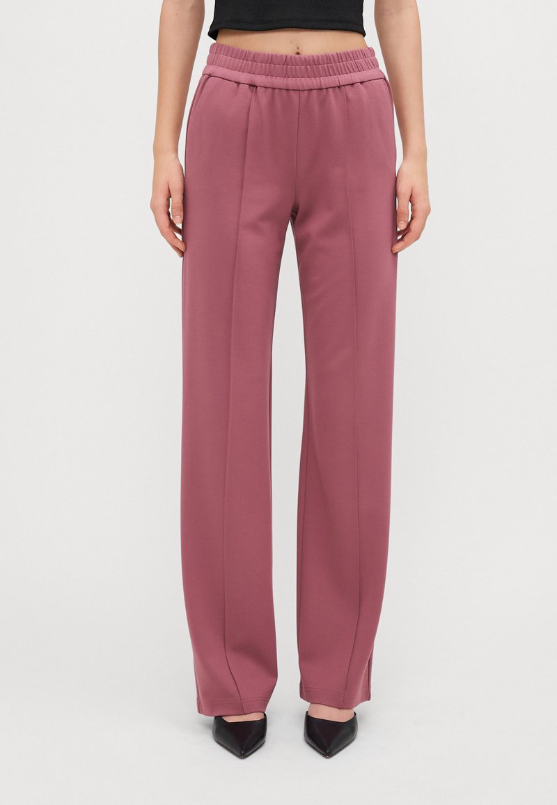 Roze, wijde broek met elastische tailleband. Gladde stof met twee verticale naden aan de voorkant, gecombineerd met zwarte puntige hakken.
