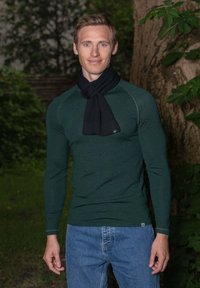 Haut à manches longues vert avec texture côtelée et écharpe noire, associé à un jean en denim bleu. Fond naturel avec feuillage.