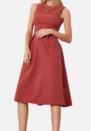 Frau trägt ein ärmelloses, rostrotes Midikleid mit tailliertem Oberteil und plissiertem Rock, accessorisiert mit einer mehrlagigen goldenen Kettenkette.