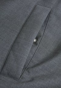 Dunkelgraue Stofftasche mit Klappe, die teilweise geöffnet ist und einen darunter liegenden metallischen Knopf zeigt.