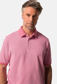 Rosa polo-tröja med klassisk krage, två knappar och korta ärmar. Tyget verkar mjukt med en subtil textur.