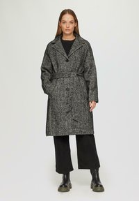 Czarny i biały płaszcz trench w wzór houndstooth z paskiem, dużymi klapami i trzema guzikami, w połączeniu z szerokimi czarnymi spodniami i masywnymi czarnymi butami.