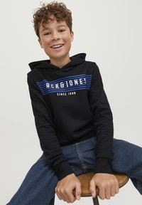 Sudadera negra con capucha y franjas horizontales blancas y azules que presentan el texto "ACK&JONES". Usada con jeans de mezclilla azules, sentado en un taburete de madera.
