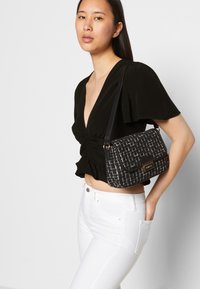 Bolsa de ombro em tweed preto e branco com fecho de aba, acabamento texturizado e detalhes em ouro. Usada com uma blusa preta e jeans brancos.
