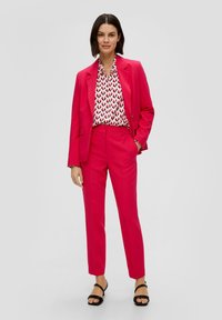 Costume rose vif comprenant une veste ajustée et un pantalon, superposé sur un chemisier blanc à motifs avec des accents noirs. Porté avec des sandales noires à lanières.