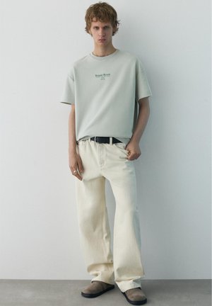 Mladý muž s krátkymi kučeravými vlasmi, oblečený v svetlosivom tričku "Botanic Resort", voľné krémové nohavice, čierny opasok a hnedé papuče, stojí proti obyčajnej stene.