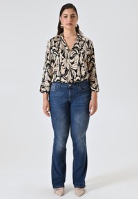 Blouse à motifs noir et crème avec des manches longues, associée à un jean droit bleu. Le modèle porte des ballerines nude pointues et des accessoires simples.