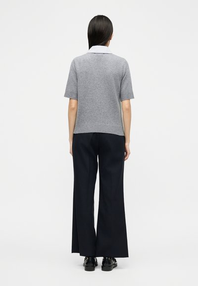 Femme aux cheveux longs et foncés portant un pull gris à manches courtes sur une chemise rayée, un pantalon large noir et des chaussures habill ées noires, debout de dos.