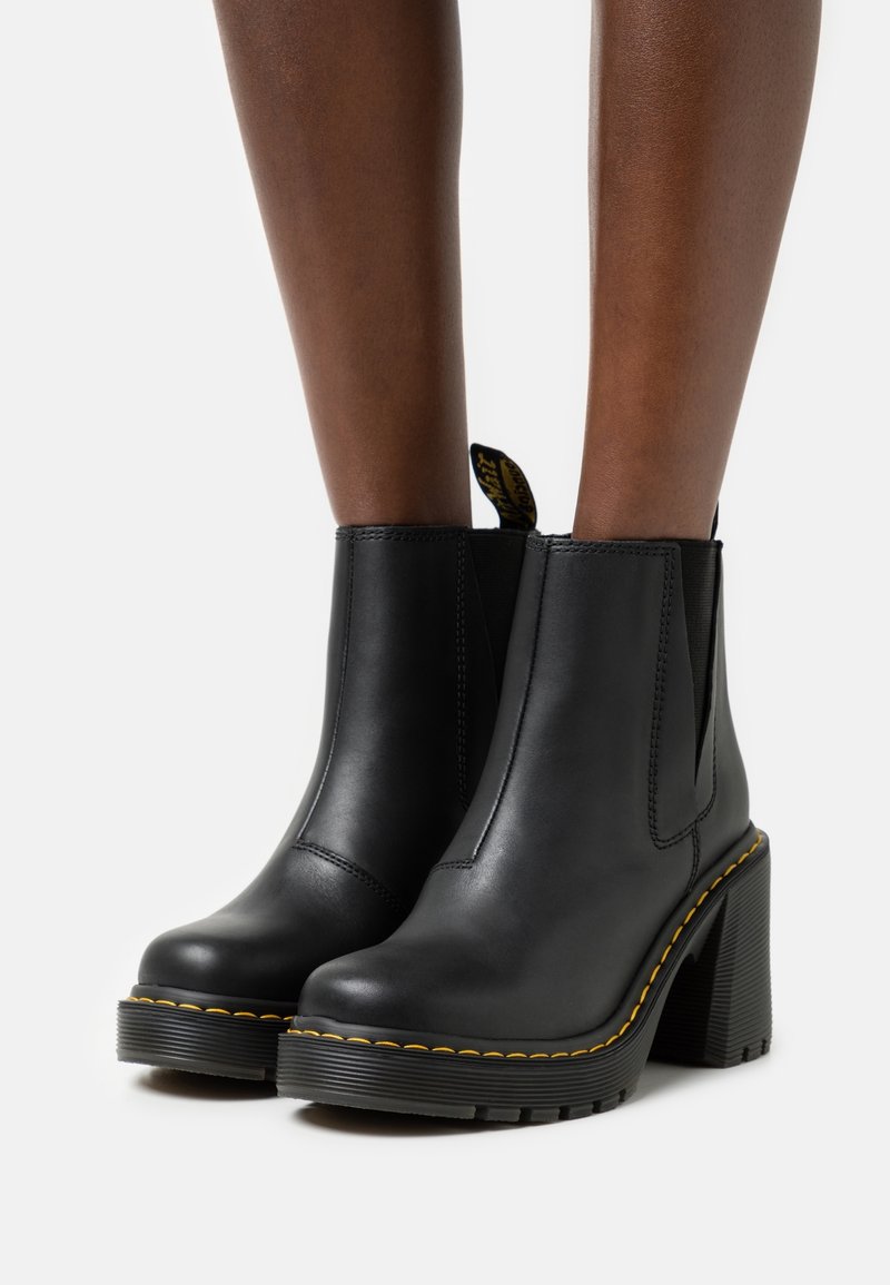 Bottes chevilles en cuir noir avec talons épaisses, surpiqûres jaunes et languettes, portées sur des jambes nues sur fond blanc.