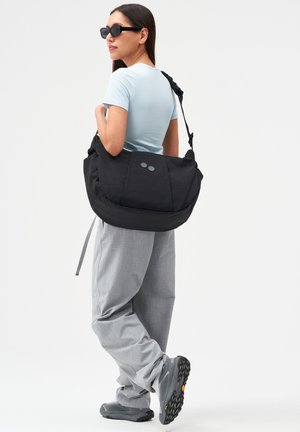 Frau mit Sonnenbrille, die ein hellblaues T-Shirt, graue Hose und graue Sneaker trägt und eine große schwarze Umhängetasche auf dem Rücken trägt.