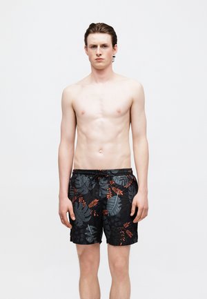 Νέος άνδρας με τα μαλλιά του χτενισμένα προς τα πίσω, φορώντας μαύρο μαγιό με γκρι και πορτοκαλί τροπικά φύλλα, στέκεται μπροστά σε απλό λευκό φόντο.