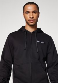 Schwarze Zip-Hoodie aus einer Baumwoll/Polyester-Mischung, mit vorderen Taschen, verstellbarem Kordelzugkapuze und gesticktem weißen Logo auf der Brust.