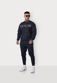 Dunkelblauer Sweatshirt und Jogginghose mit weißem "ESTEEM"-Logo, runder Halsausschnitt und gerippten Bündchen. Getragen mit weißen Sneakers und Sonnenbrille.