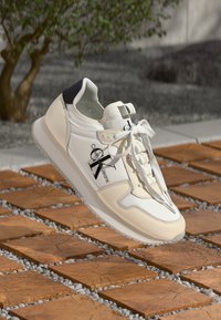 Tenis alb cu un superior din piele netedă, accente negre, design cu șireturi, detalii cu logo și talpă din cauciuc. Așezat pe un fundal texturat.