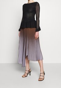 Kobieta w czarnej przezroczystej bluzce z długim rękawem oraz w brązowo-liliowej spódnicy midi w fałdy z ombre, nosząca czarne sandały na obcasie z paskami.