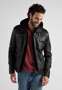 Zwarte leren jas met een afneembare capuchon, voorzien van ritszakken, een aansluitend ontwerp en ribbeltextuur op de mouwen. Draagt over een rood shirt.