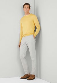 Suéter de punto amarillo, cuello blanco, pantalones gris claro y zapatos de gamuza marrón. El modelo se encuentra de pie contra un fondo gris claro, con las manos en los bolsillos.