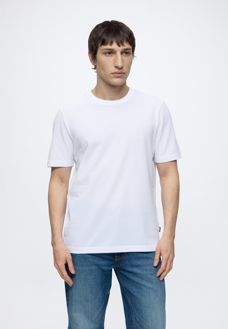 Junger Mann steht still, trägt ein einfaches weißes kurzärmeliges Hemd und blaue Jeans vor weißem Hintergrund.