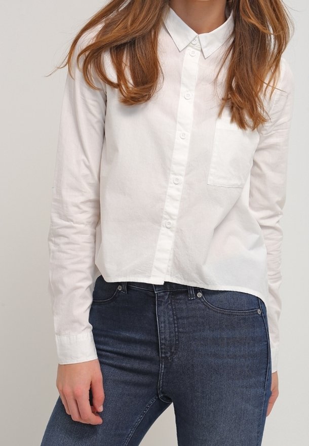 Femme portant une chemise blanche à boutons avec manches longues et un jean taille haute bleu foncé, debout devant un fond uni.
