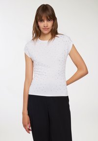 Biała bluzka z krótkim rękawem o dopasowanym kroju, ozdobiona małymi srebrnymi ćwiekami ułożonymi w wzór. Gładki, elastyczny materiał.