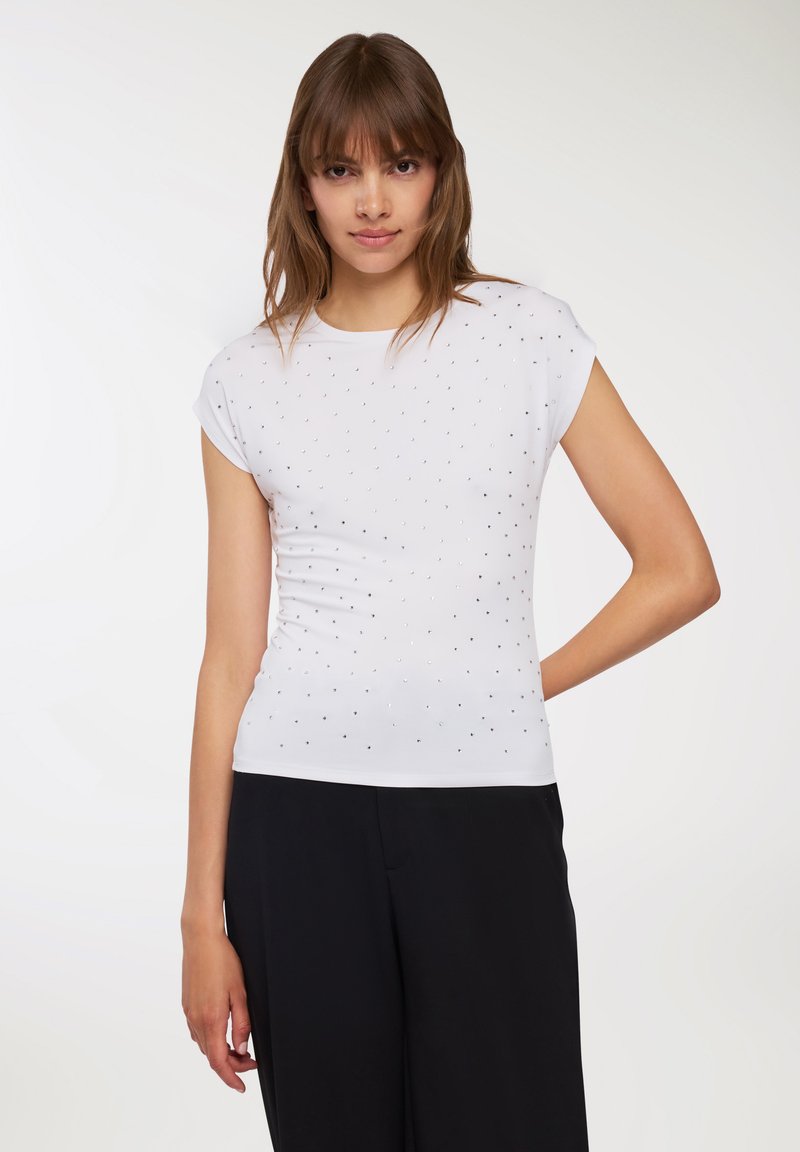 Biała bluzka z krótkim rękawem o dopasowanym kroju, ozdobiona małymi srebrnymi ćwiekami ułożonymi w wzór. Gładki, elastyczny materiał.