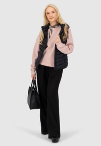Zwarte puffer vest over een lichtroze sweatshirt, gecombineerd met zwarte wijde broek en een zwarte handtas. Lang haar, casual stijl.