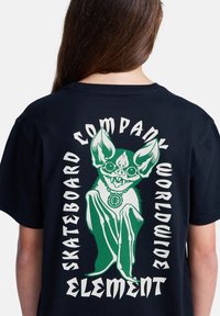 Persona che indossa una maglietta nera con un pipistrello stilizzato verde e bianco con grandi orecchie e la scritta "SKATEBOARD COMPANY WORLDWIDE ELEMENT" sulla schiena.