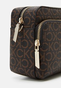 Zbliżenie na ciemnobrązową saszetkę Calvin Klein z powtarzającym się wzorem logo CK i złotymi suwakami w dwóch komorach.