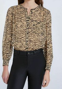 Beige Bluse mit schwarzen Tigermustern, geknöpfter Front, rundem Ausschnitt und einer Brusttasche, kombiniert mit schwarzen, figurbetonten Hose. Strukturierter Stoff.