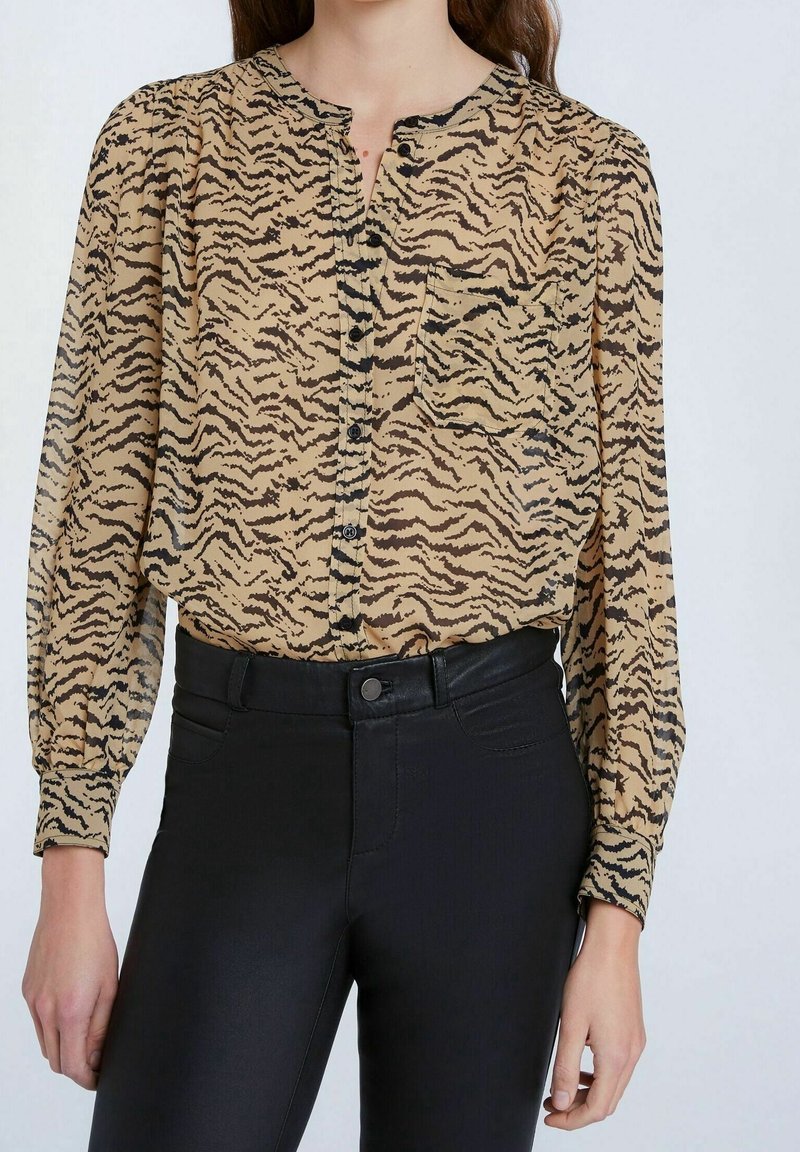 Blusa beige con strisce di tigre nere, chiusura frontale a bottoni, scollo rotondo e tasca sul petto, abbinata a pantaloni neri aderenti. Tessuto a trama.